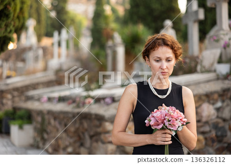 Sad woman in black dress with bouquet of flowers and handkerchief in hands, stands in cemetery 136326112