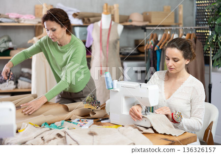 Female tailor sews on a sewing machine while another tailor cuts out patterns for dress with scissors Female tailor sews on a sewing machine while another tailor cuts out patterns for dress with scissors 136326157