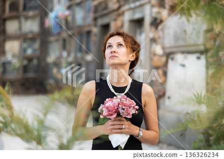 Upset woman in black dress, who lost loved one, brings flowers to grave 136326234