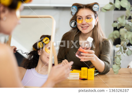 Woman in colorful curlers applying under eye patches by mirror with bestie Woman in colorful curlers applying under eye patches by mirror with bestie 136326287
