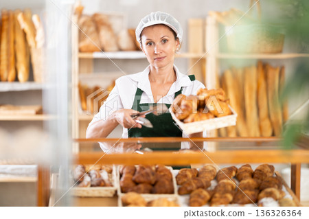 Woman employee puts croissants in window, arranges display of goods at bakery. Woman employee puts croissants in window, arranges display of goods at bakery. 136326364