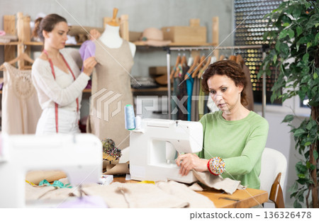 Woman tailor at work in cloth design shop. Companion work in background with mannequin 136326478