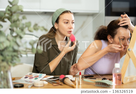 Two female friends applying makeup by vanity mirror at home Two female friends applying makeup by vanity mirror at home 136326519