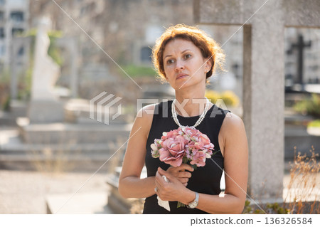 Grieving woman in black with flowers standing in cemetery Grieving woman in black with flowers standing in cemetery 136326584