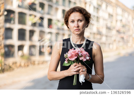 Sorrowful woman in black standing in cemetery with flowers Sorrowful woman in black standing in cemetery with flowers 136326596