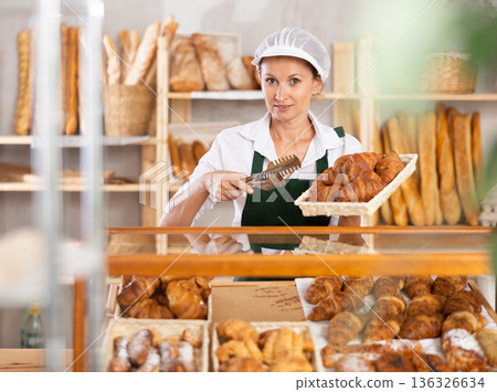 Woman employee puts croissants in window, arranges display of goods at bakery. 136326634