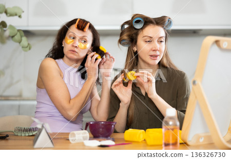 Two adult besties applying hair curlers in front of tabletop mirror Two adult besties applying hair curlers in front of tabletop mirror 136326730