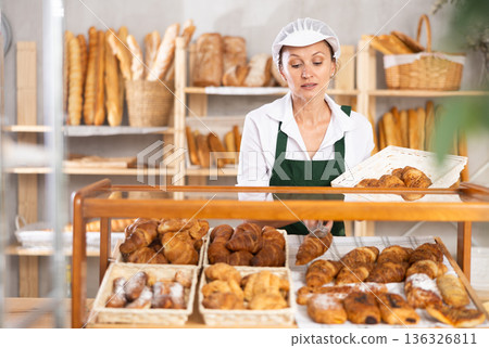 Woman employee puts croissants in window, arranges display of goods at bakery. 136326811