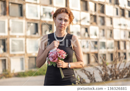 Sad woman in black dress with bouquet of flowers and handkerchief in hands, stands in cemetery 136326881
