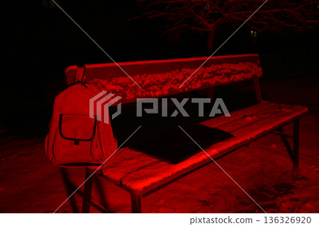 backpack hanging on the back of a snowy bench in red light 136326920