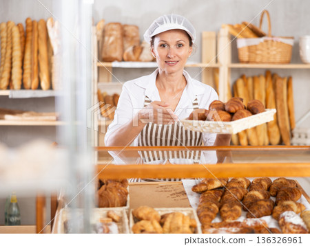 Positive female bakery seller with tongs in his hands offers to buy fresh croissants 136326961