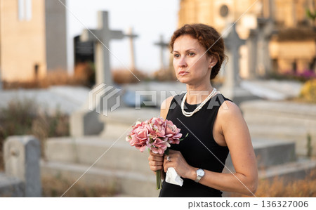 Sad woman in black dress with bouquet of flowers and handkerchief in hands, stands in cemetery Sad woman in black dress with bouquet of flowers and handkerchief in hands, stands in cemetery 136327006