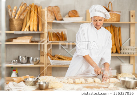 Woman works in bakery as baker, cuts dough into portions, forms pieces of dough to create buns. 136327036