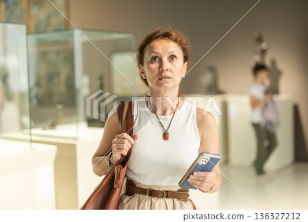 Woman with phone looking around in museum hall Woman with phone looking around in museum hall 136327212
