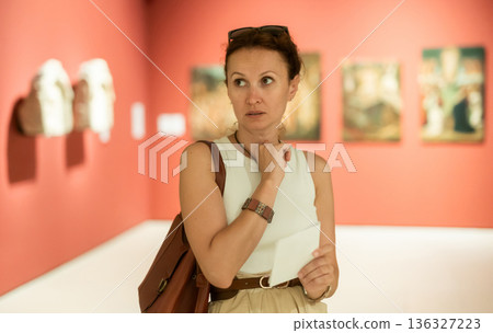 Portrait of pretty young woman visitor with paper guide at exhibition of paintings in museum Portrait of pretty young woman visitor with paper guide at exhibition of paintings in museum 136327223