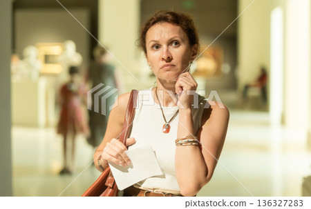 Thoughtful woman visiting museum exhibition of statues with information booklet in hand 136327283