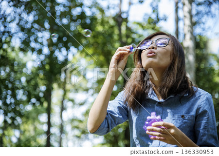 Adorable happy cute girl playful foam bubbles in green playground in summer outdoors. Funny Cheerful girl in the park happiness times. Bubbles blowing soap playful in nature park. 136330953