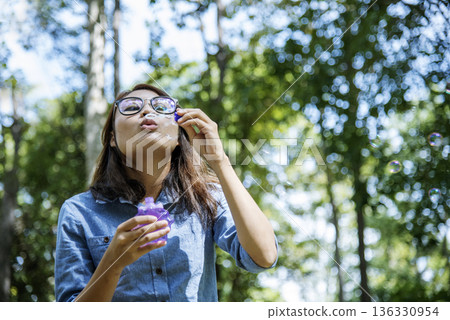 Adorable happy cute girl playful foam bubbles in green playground in summer outdoors. Funny Cheerful girl in the park happiness times. Bubbles blowing soap playful in nature park. 136330954