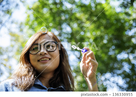 Adorable happy cute girl playful foam bubbles in green playground in summer outdoors. Funny Cheerful girl in the park happiness times. Bubbles blowing soap playful in nature park. 136330955
