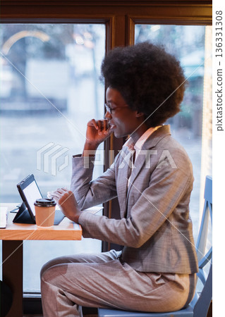 A professional woman with an afro hairstyle works on her tablet while sitting at a table in a cafe with a coffee cup nearby A professional woman with an afro hairstyle works on her tablet while sitting at a table in a cafe with a coffee cup nearby 136331308