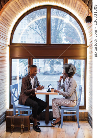 Two professionals in business attire engage in a conversation over coffee and a tablet at a table in a modern cafe with a large arched window Two professionals in business attire engage in a conversation over coffee and a tablet at a table in a modern cafe with a large arched window 136331309