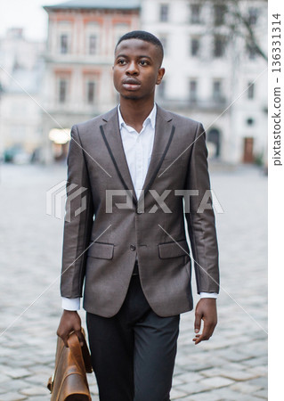A young Black man in a suit walks down a cobblestone street carrying a leather briefcase, looking thoughtfully into the distance 136331314