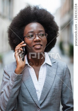 A professional Black woman with an afro hairstyle wears glasses and a grey suit while holding a smartphone to her ear and speaking 136331334