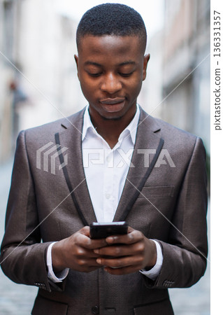 A young Black man in a formal suit looks down at his smartphone while standing outdoors on a city street A young Black man in a formal suit looks down at his smartphone while standing outdoors on a city street 136331357