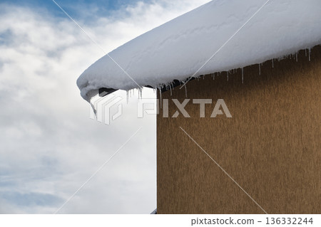 Snow-covered roof with icicles hanging against a cloudy sky 136332244
