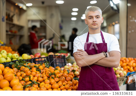 Young man seller in apron stands in trading hall in vegetable store Young man seller in apron stands in trading hall in vegetable store 136333631