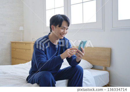 A man in his twenties sitting on a bed and using a smartphone A man in his twenties sitting on a bed and using a smartphone 136334714