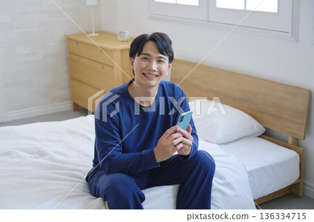 A man in his 20s sitting on a bed and holding a smartphone, looking at the camera, portrait A man in his 20s sitting on a bed and holding a smartphone, looking at the camera, portrait 136334715