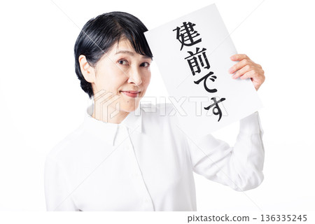一位面露得意的老婦人，一手拿著一張寫著「建前」的紙。 136335245