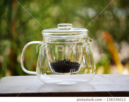Clear glass teapot with loose black tea leaves inside, placed on wooden table with natural green bokeh background. Minimal tea brewing concept, calm lifestyle, healthy drink, clean design, relaxation 136336012