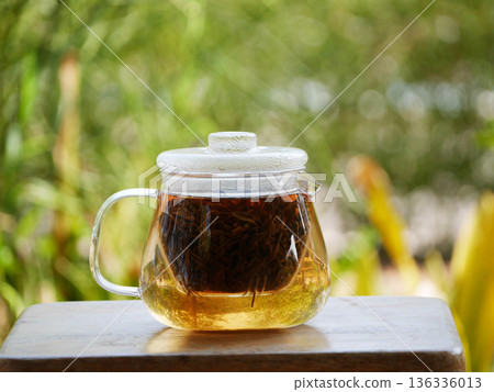 glass teapot brewing loose black tea with hot water, golden tea color visible inside. Minimal tea preparation scene on wooden table with natural green bokeh background, calm lifestyle, healthy drink 136336013