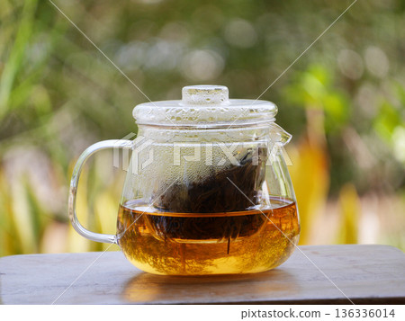 glass teapot brewing loose black tea with hot water, golden tea color visible inside. Minimal tea preparation scene on wooden table with natural green bokeh background, calm lifestyle, healthy drink 136336014