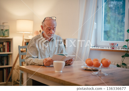 Older man reading book i kitchen. 136336677