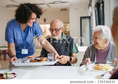 Caregiver serving lunch to elderly residents in a care home. Caregiver serving lunch to elderly residents in a care home. 136336832