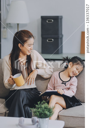 A cheerful Asian mother helps her young daughter do homework and color a book at home, while the father works on a laptop in the background. A cozy, bright, and modern family lifestyle. A cheerful Asian mother helps her young daughter do homework and color a book at home, while the father works on a laptop in the background. A cozy, bright, and modern family lifestyle. 136337017