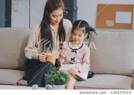 A cheerful Asian mother helps her young daughter do homework and color a book at home, while the father works on a laptop in the background. A cozy, bright, and modern family lifestyle. A cheerful Asian mother helps her young daughter do homework and color a book at home, while the father works on a laptop in the background. A cozy, bright, and modern family lifestyle. 136337019