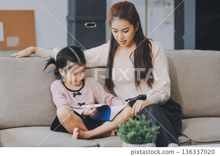 A cheerful Asian mother helps her young daughter do homework and color a book at home, while the father works on a laptop in the background. A cozy, bright, and modern family lifestyle. A cheerful Asian mother helps her young daughter do homework and color a book at home, while the father works on a laptop in the background. A cozy, bright, and modern family lifestyle. 136337020