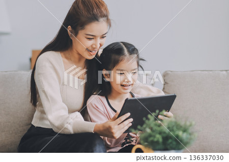 A cheerful Asian mother helps her young daughter do homework and color a book at home, while the father works on a laptop in the background. A cozy, bright, and modern family lifestyle. A cheerful Asian mother helps her young daughter do homework and color a book at home, while the father works on a laptop in the background. A cozy, bright, and modern family lifestyle. 136337030