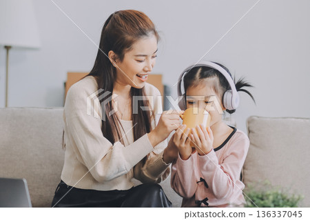 A cheerful Asian mother helps her young daughter do homework and color a book at home, while the father works on a laptop in the background. A cozy, bright, and modern family lifestyle. A cheerful Asian mother helps her young daughter do homework and color a book at home, while the father works on a laptop in the background. A cozy, bright, and modern family lifestyle. 136337045