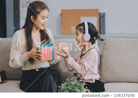 A cheerful Asian mother helps her young daughter do homework and color a book at home, while the father works on a laptop in the background. A cozy, bright, and modern family lifestyle. 136337056