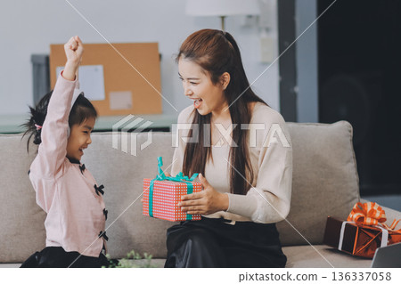A cheerful Asian mother helps her young daughter do homework and color a book at home, while the father works on a laptop in the background. A cozy, bright, and modern family lifestyle. A cheerful Asian mother helps her young daughter do homework and color a book at home, while the father works on a laptop in the background. A cozy, bright, and modern family lifestyle. 136337058