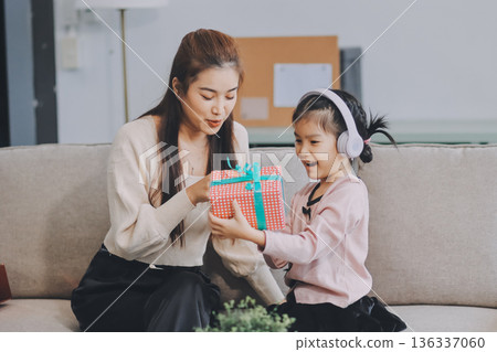 A cheerful Asian mother helps her young daughter do homework and color a book at home, while the father works on a laptop in the background. A cozy, bright, and modern family lifestyle. 136337060