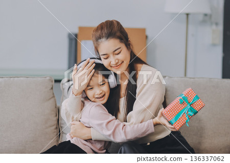 A cheerful Asian mother helps her young daughter do homework and color a book at home, while the father works on a laptop in the background. A cozy, bright, and modern family lifestyle. A cheerful Asian mother helps her young daughter do homework and color a book at home, while the father works on a laptop in the background. A cozy, bright, and modern family lifestyle. 136337062