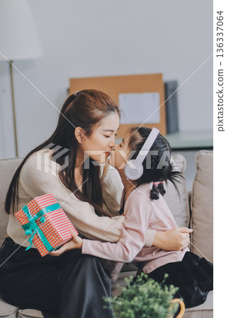 A cheerful Asian mother helps her young daughter do homework and color a book at home, while the father works on a laptop in the background. A cozy, bright, and modern family lifestyle. 136337064
