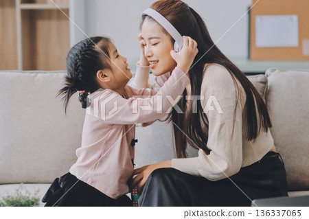 A cheerful Asian mother helps her young daughter do homework and color a book at home, while the father works on a laptop in the background. A cozy, bright, and modern family lifestyle. A cheerful Asian mother helps her young daughter do homework and color a book at home, while the father works on a laptop in the background. A cozy, bright, and modern family lifestyle. 136337065
