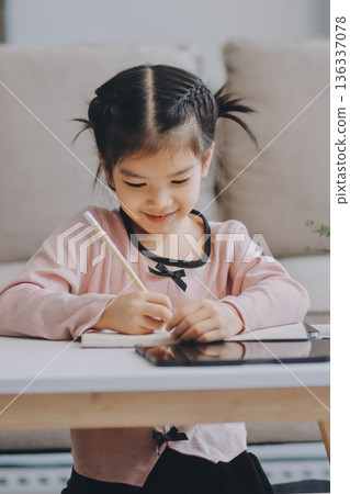 A cheerful Asian mother helps her young daughter do homework and color a book at home, while the father works on a laptop in the background. A cozy, bright, and modern family lifestyle. A cheerful Asian mother helps her young daughter do homework and color a book at home, while the father works on a laptop in the background. A cozy, bright, and modern family lifestyle. 136337078